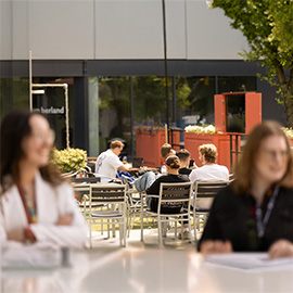 Two people sat at a table, with a group of students in the background sat outside at habita, the students union bar