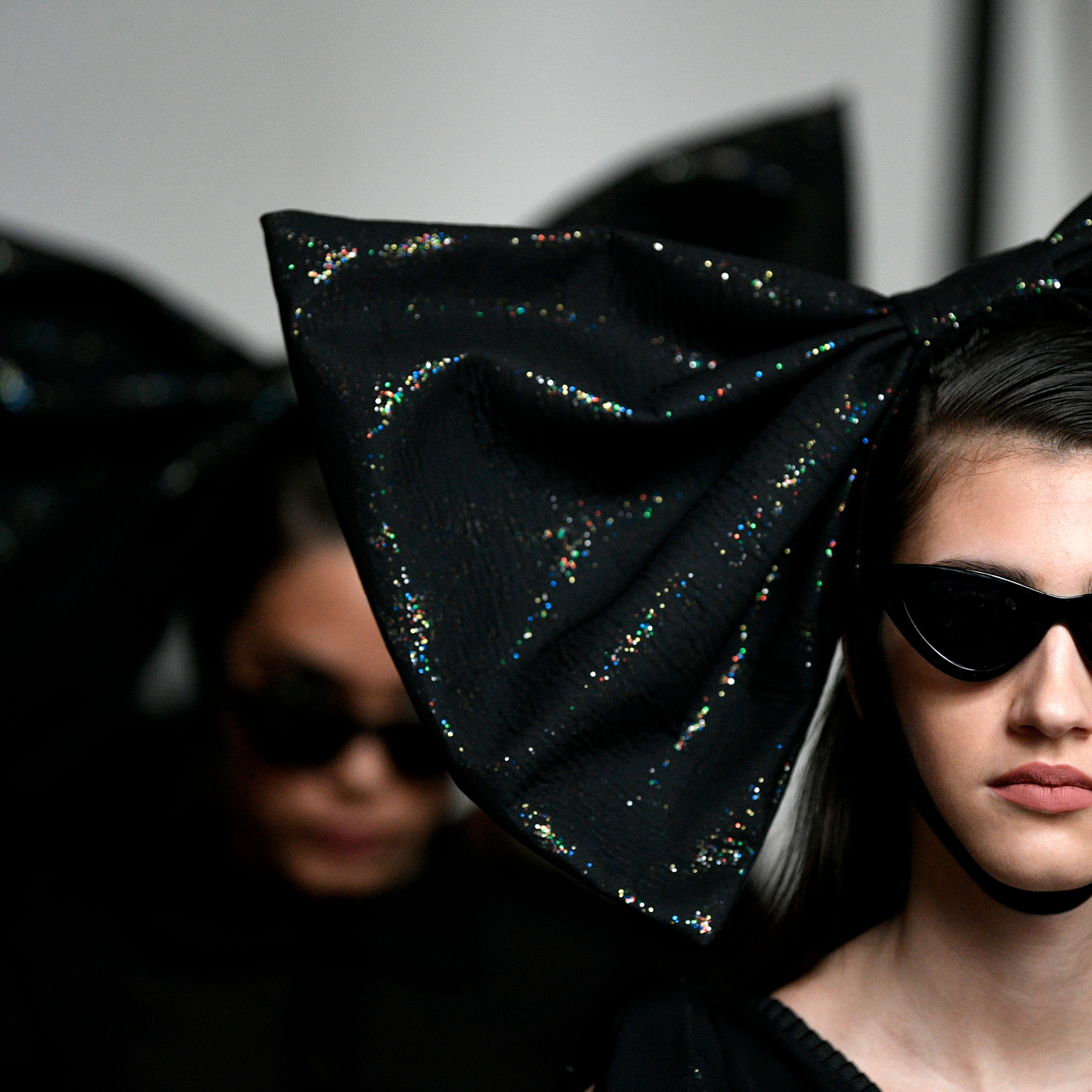 close up of a girl on a catwalk wearing black sunglasses and an oversized black glittery bow on her head