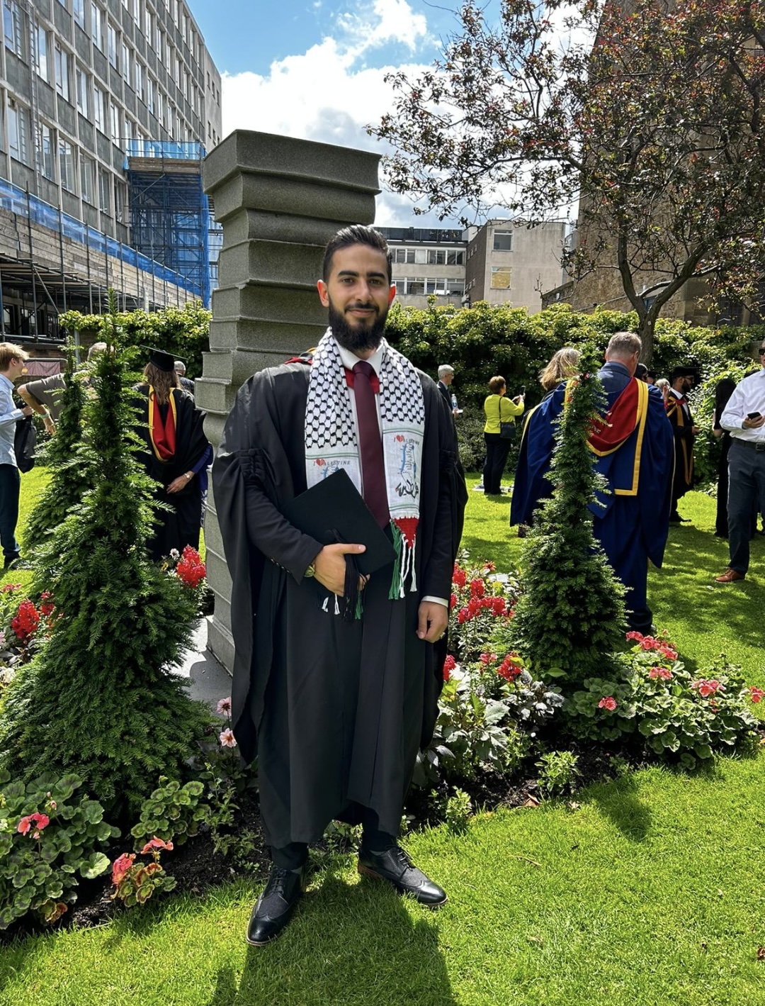 Man wearing a graduation gown holding a graduation cap in his hands. He is standing in front of a statue of books having his picture taken. 