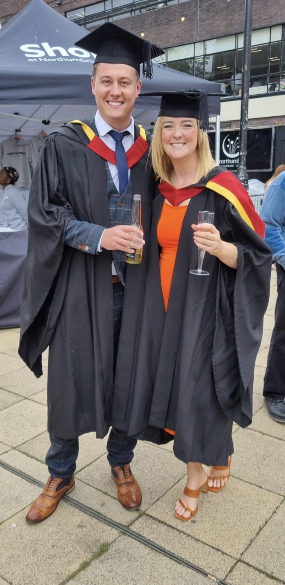 Man and woman wearing graduation gowns and caps smiling at the camera while holding a drink