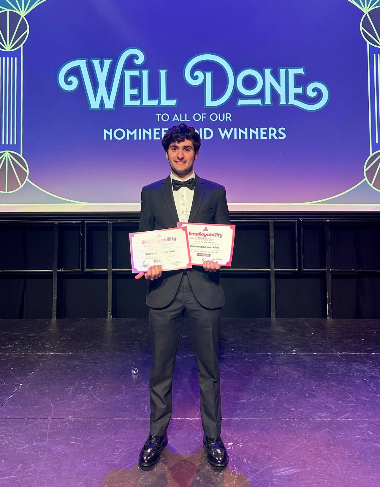 Image of male student standing holding 2 awards certificates smiling at the camera