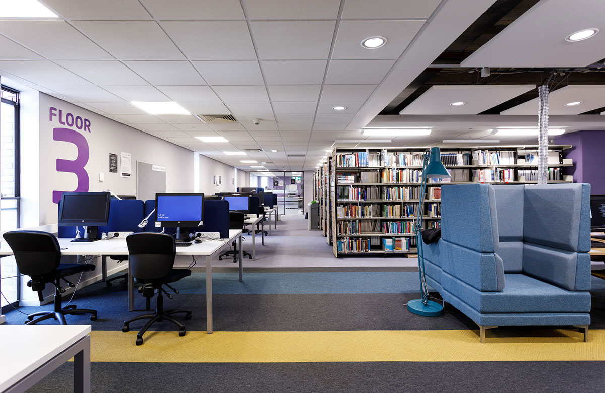 An image of the university library showing shelves with books, some comfortable chairs with floor lamps, desks and computers to the left and a wall sign saying Floor 3