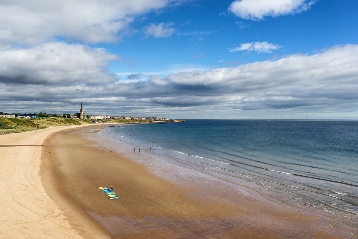 An image of Longsands Beach at Tynemouth, UK. Showing blue skies with white fluffy clouds, small waves and some colourful surfboards laid out on the sand