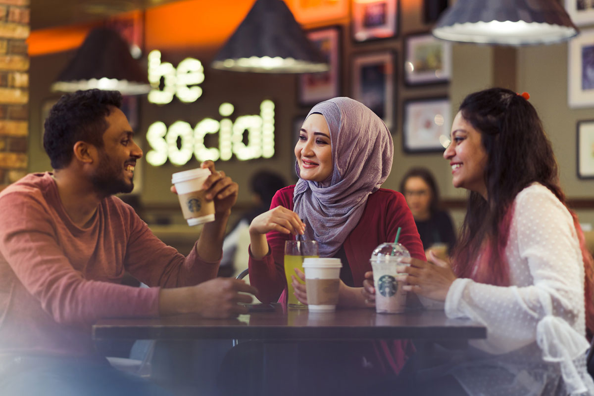 An image of three students round a table in the students union. a male student on the left is smiling at two female students, there are coffee cups on the table and a light up sign in the background says be social