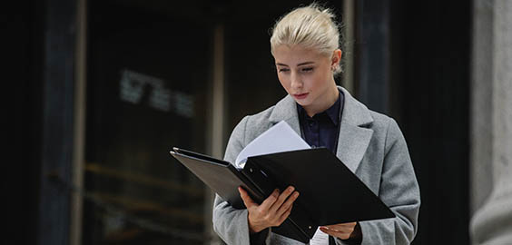 A woman dressed in a smart grey coat looking at an open black binder with documents
