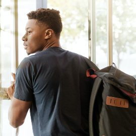 Black young man looking out a window; carrying a black-brown backpack