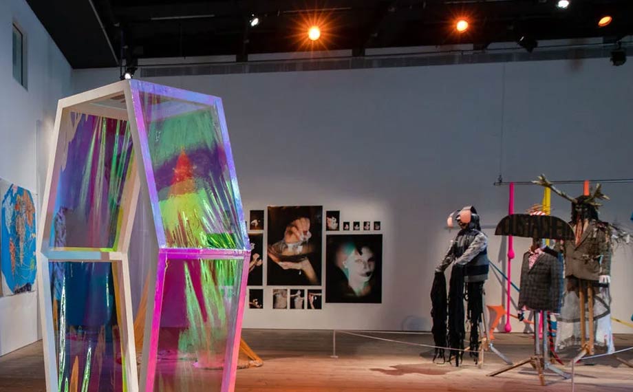 Image of an art exhibition space. White walls with wood flooring. to the left we have a coffin shaped structure with see through rainbow coloured covering. On the back wall are some photographs and to the right some mannequins.