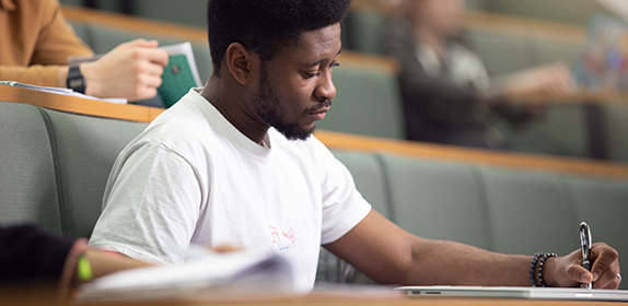 Close-up of a student making notes in a lecture theatre. He is wearing a white t-shirt and holding a pen.