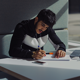 Close-up of a male student sat in a pod, making notes in a notepad with his laptop also open.