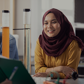 Close-up of a student in a maroon headscarf and yellow top leaning on a bench - she is smiling as someone shows her notes.