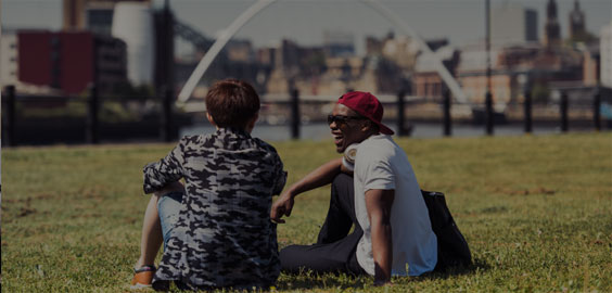 Image: Two make students sitting on the grass talking with the Newcastle Quayside scenery in the background.