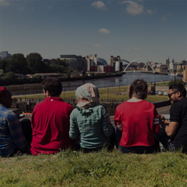 Male and female international students sitting on the grass with their backs to the camera, with the Quayside in the foreground.