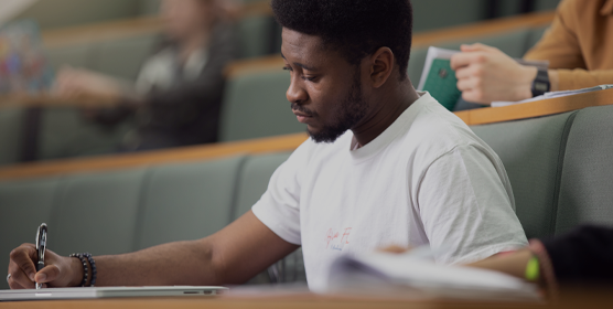 An international student wearing a white t-shirt is making notes during a lecture