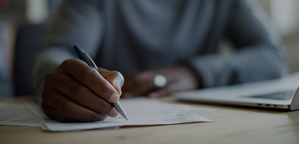 Close-up of an individual writing on paper. They're sat at a desk with a laptop open next to them.