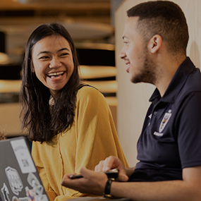 Close-up of two students sat in Student Central, laughing. They are facing one another, one is wearing a blue t-shirt and has an open laptop on their knees. The other is wearing a yellow blouse.