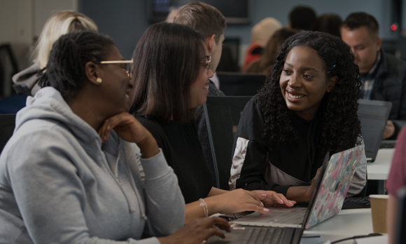 Three international students are gathered around a laptop on campus, they are smiling and talking.