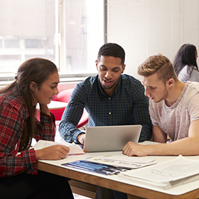 Three students working collaboratively. They're gathered around a laptop.