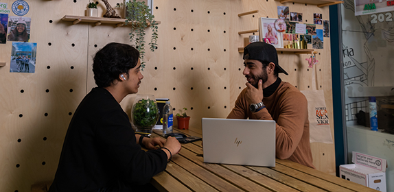 Two students sat at a table in the Students' Union. They have a laptop open between them and are talking.