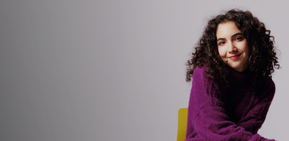Image of a female student with curly hear sat on a yellow chair, against a white background as part of the 'I Am Northumbria' campaign.