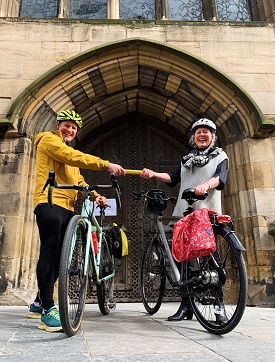 Image shows two people on bikes outside Newcastle cathedral. One is handing a relay baton to the other.