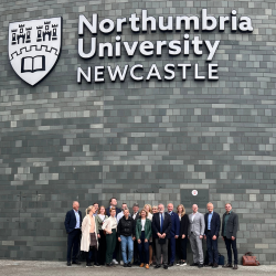 A group of people pictured standing in front of a grey brick wall with a large Northumbria University logo in the background