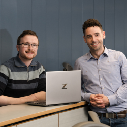 two men pictured at a lecture theatre seating area with a laptop sat on the desk between them