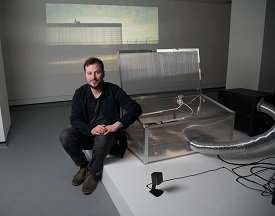 Pictured is artist Paul Dolan sitting in front of an installation with an image projected onto the wall behind him