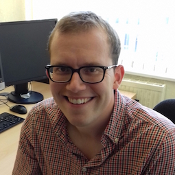 a man sitting at a desk with a laptop and smiling at the camera