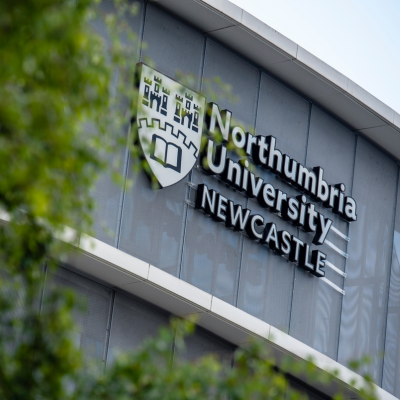 Northumbria University sign on the exterior of a grey building with green foliage framing the photograph. 