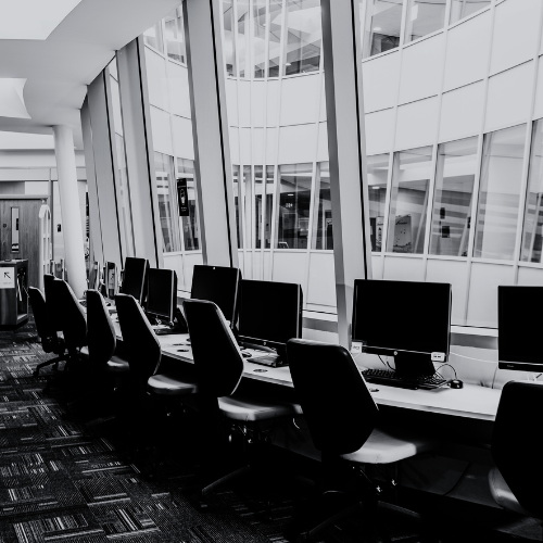 Black-and-white photo of a row of empty desktop computers with black chairs along a curved desk, set beneath tall floor-to-ceiling windows 