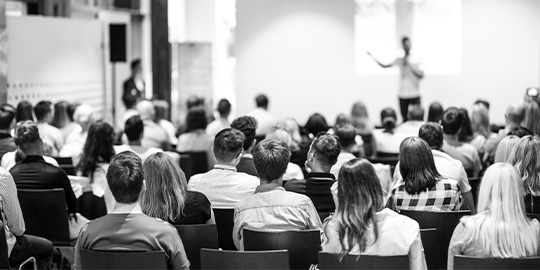 Image of a lecture theatre taken from the behind the audience with a speaker in the distance. 