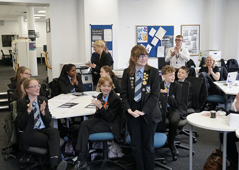 Seated children in a classroom applauding female student who is standing