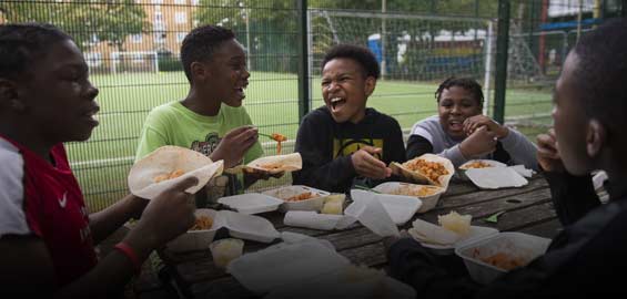 Five children sit round a table eating food on a park bench. Behind them is a five-a-side football court.
