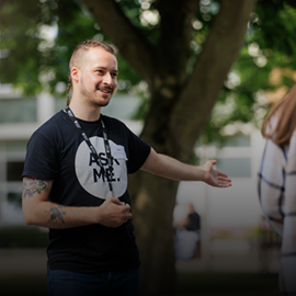 A man wearing a black T-Shirt that says 'ask me' in white. He is holding out an arm to help direct a student.