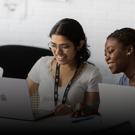 Two female students sat at a desk smiling towards a laptop