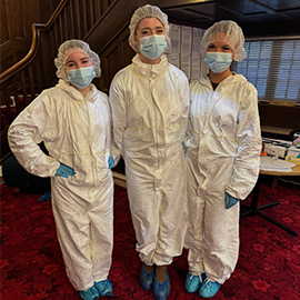 Three people wearing white protective suits, hairnets, face masks, gloves, and blue shoe covers standing on a red carpeted floor indoors.