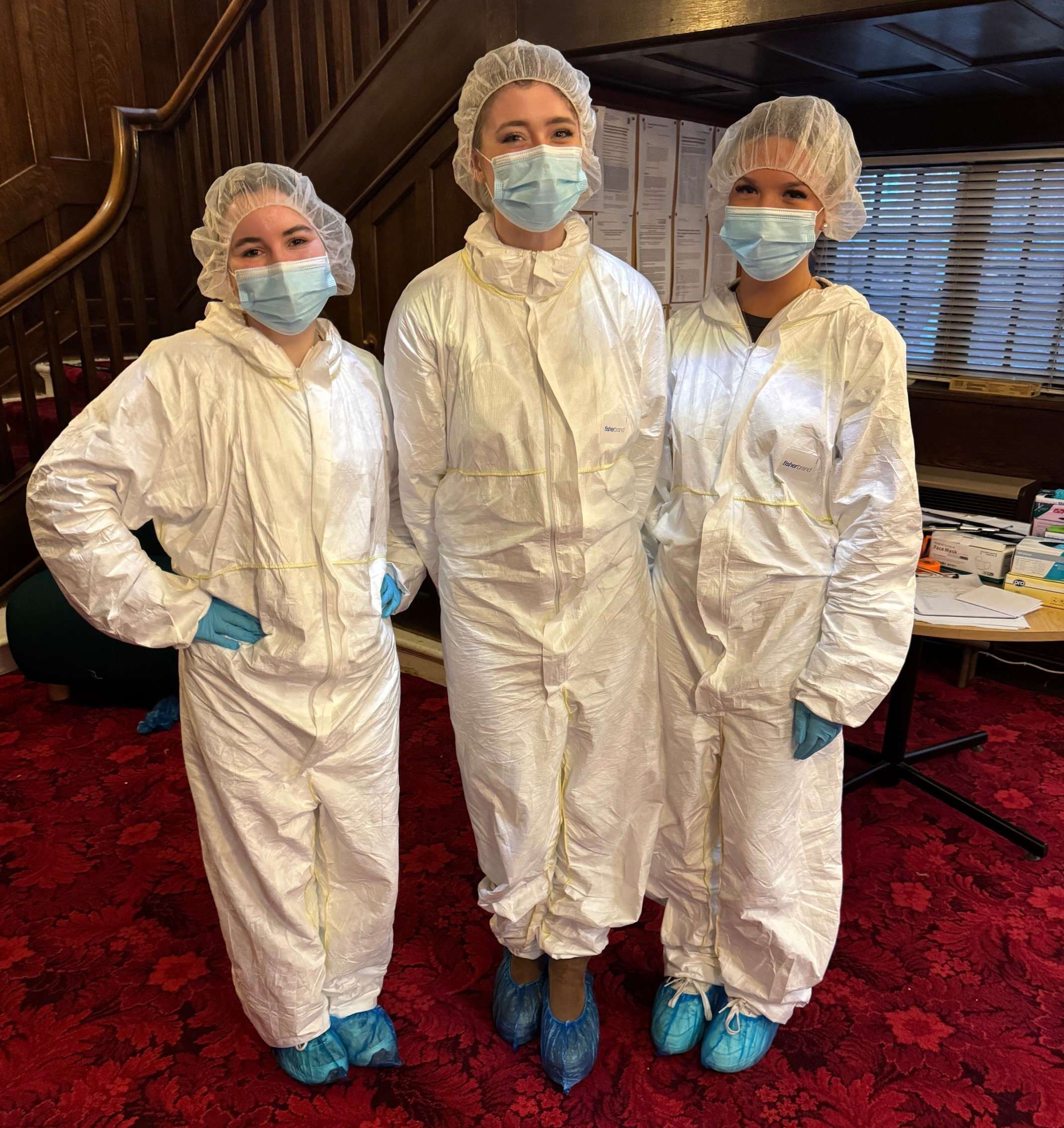 Three people wearing white protective suits, hairnets, face masks, gloves, and blue shoe covers standing on a red carpeted floor indoors.
