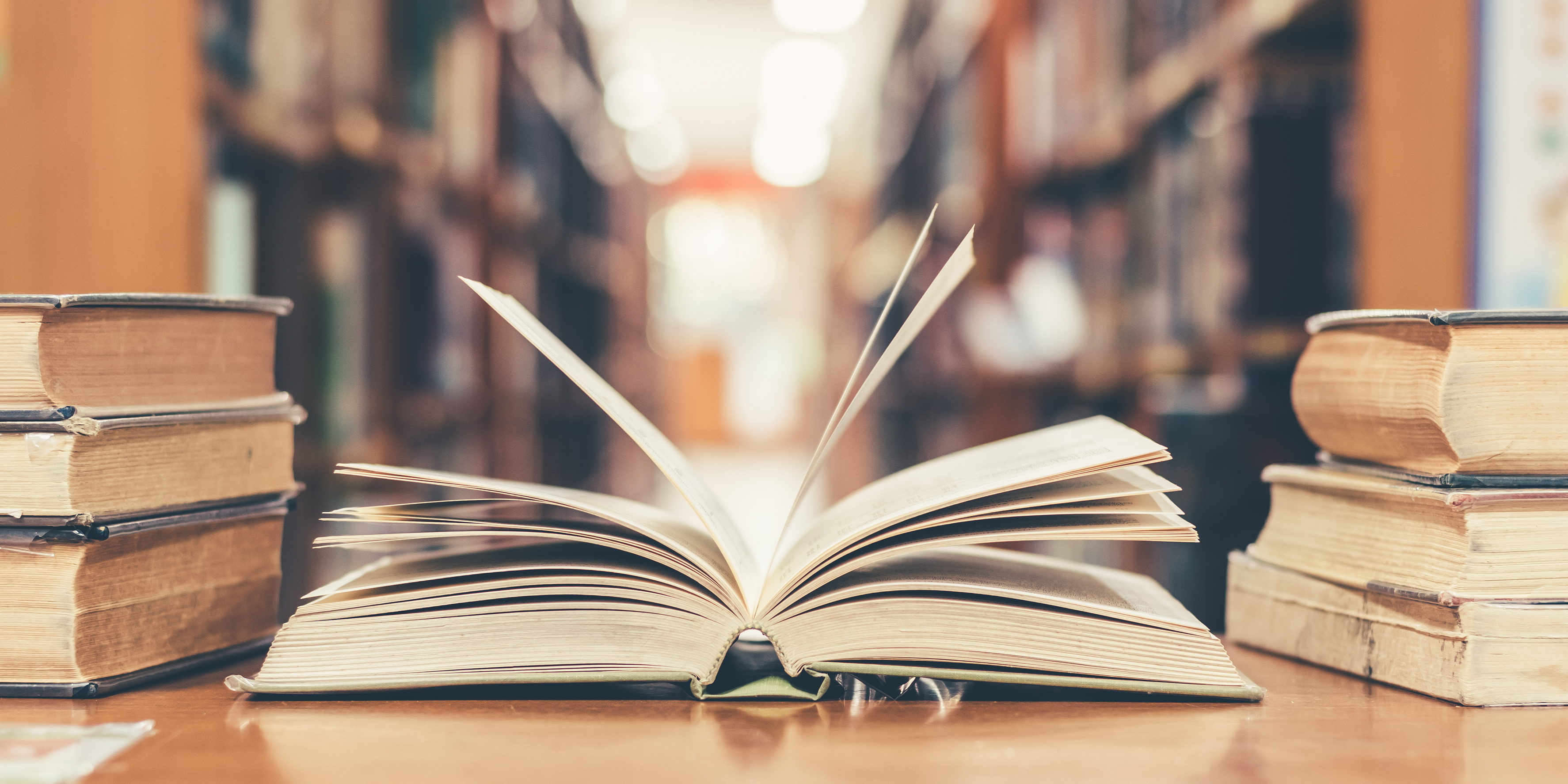 Two stacks of books on a desk with an open book in the centre. In the background there out of focus bookshelves.
