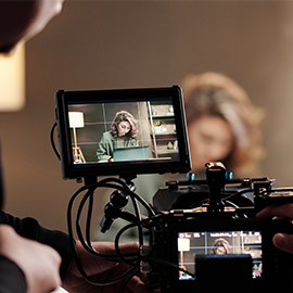 Camera monitor showing a woman working on a laptop during a video shoot, with the camera crew adjusting equipment