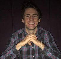 A man with black hair smiling. He is in a checked shirt with his elbows on a table with one hand on top of the other.