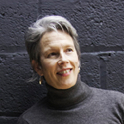 A woman with short grey hair wearing a grey turtleneck jumper. She is standing in front of a dark grey brick wall. 