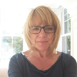 A woman with short blond hair and a fringe. She is wearing black rimmed rectangular glasses.