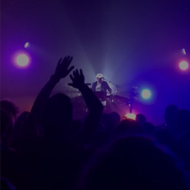 some people at a gig dancing to a dj. Once person's hand is in the air while they dance. The image has purple and blue tones of colour.