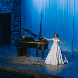 a woman on stage wearing a ball gown. a man plays piano beside her.