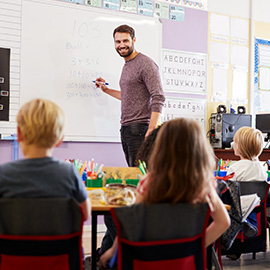 Male Teacher Standing At Whiteboard Teaching Maths Lesson To Elementary Pupils In School Classroom